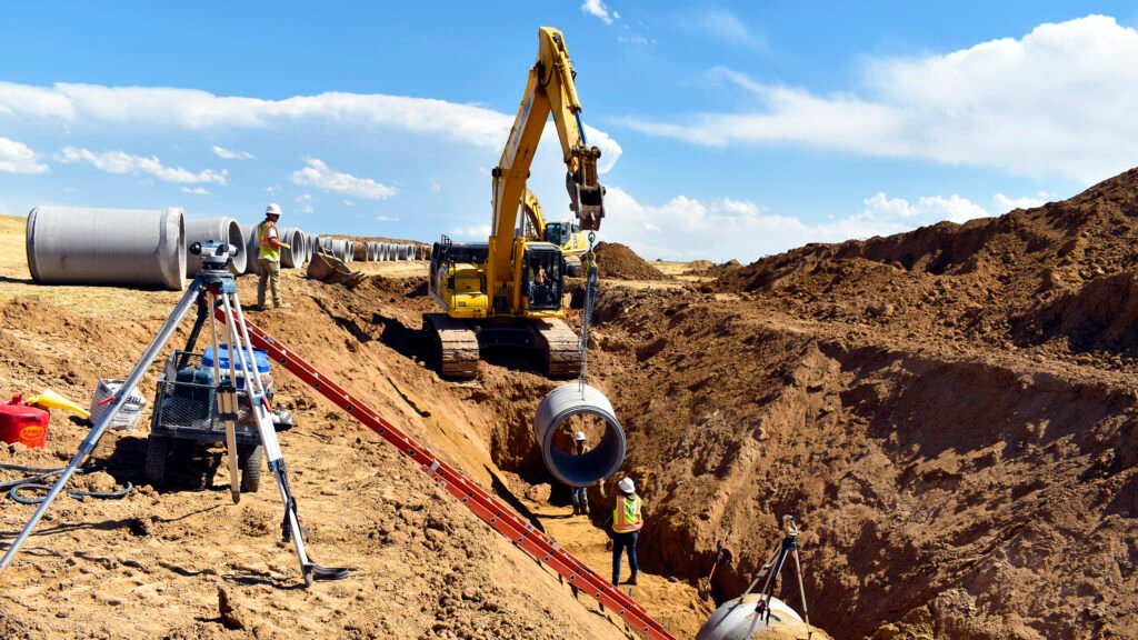 Excavatorlowering large cement pipes to workers in ditch on a job site